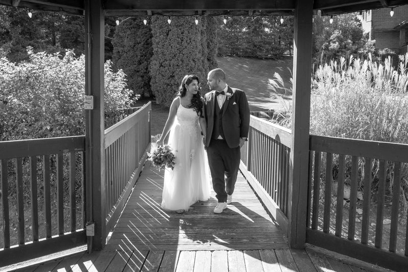 A bride and groom walk hand-in-hand on a wooden bridge in a lush garden, framed by pillars and railings under string lights.