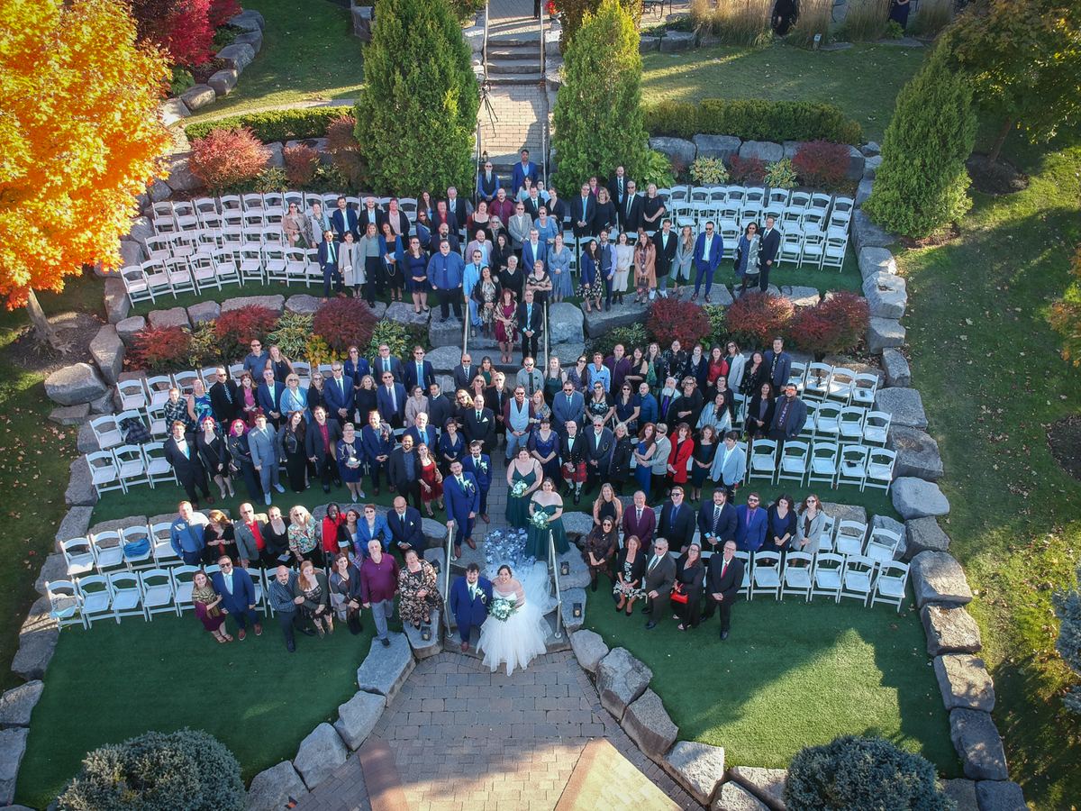 Bride and groom stand at center of stone aisle, surrounded by guests in white chairs under autumn trees.