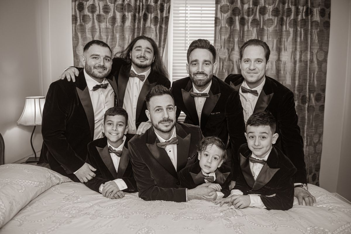 A groom and his groomsmen, including three young boys, pose in matching tuxedos on a bed, illuminated by soft light.