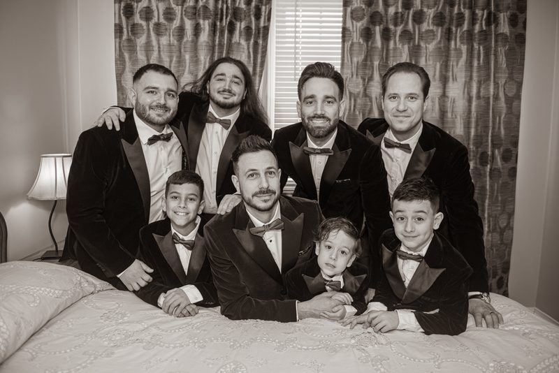 A groom and his groomsmen, including three young boys, pose in matching tuxedos on a bed, illuminated by soft light.