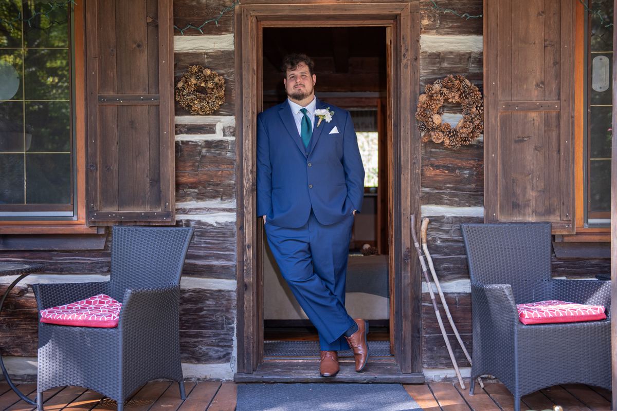 A groom in a navy suit stands confidently by the door of a rustic log cabin, hands in pockets, gazing at the camera.