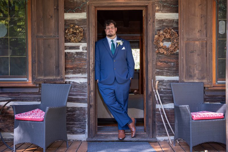 A groom in a navy suit stands confidently by the door of a rustic log cabin, hands in pockets, gazing at the camera.