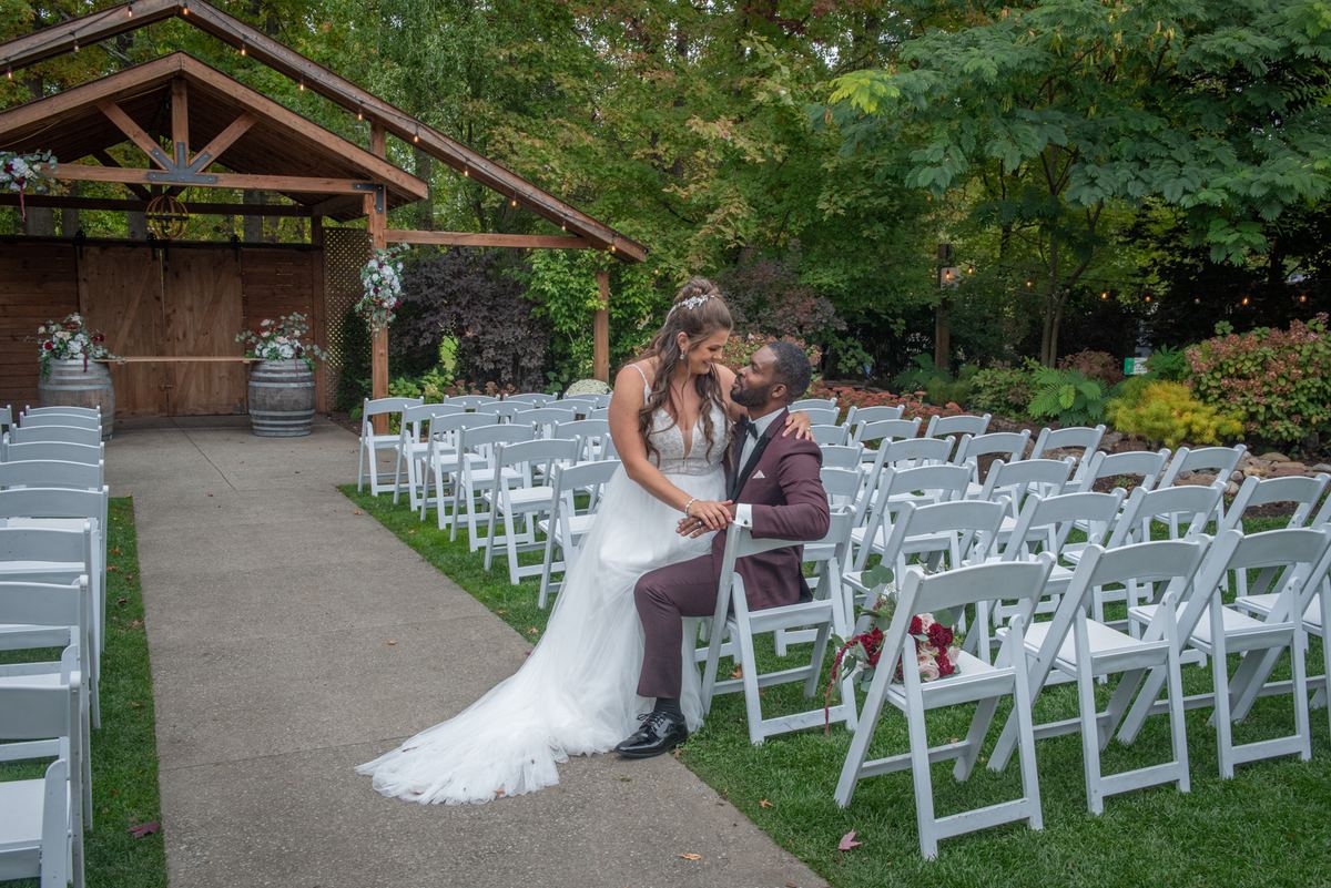A bride leans in to kiss her groom's cheek beside white chairs and lush greenery at an outdoor wedding.