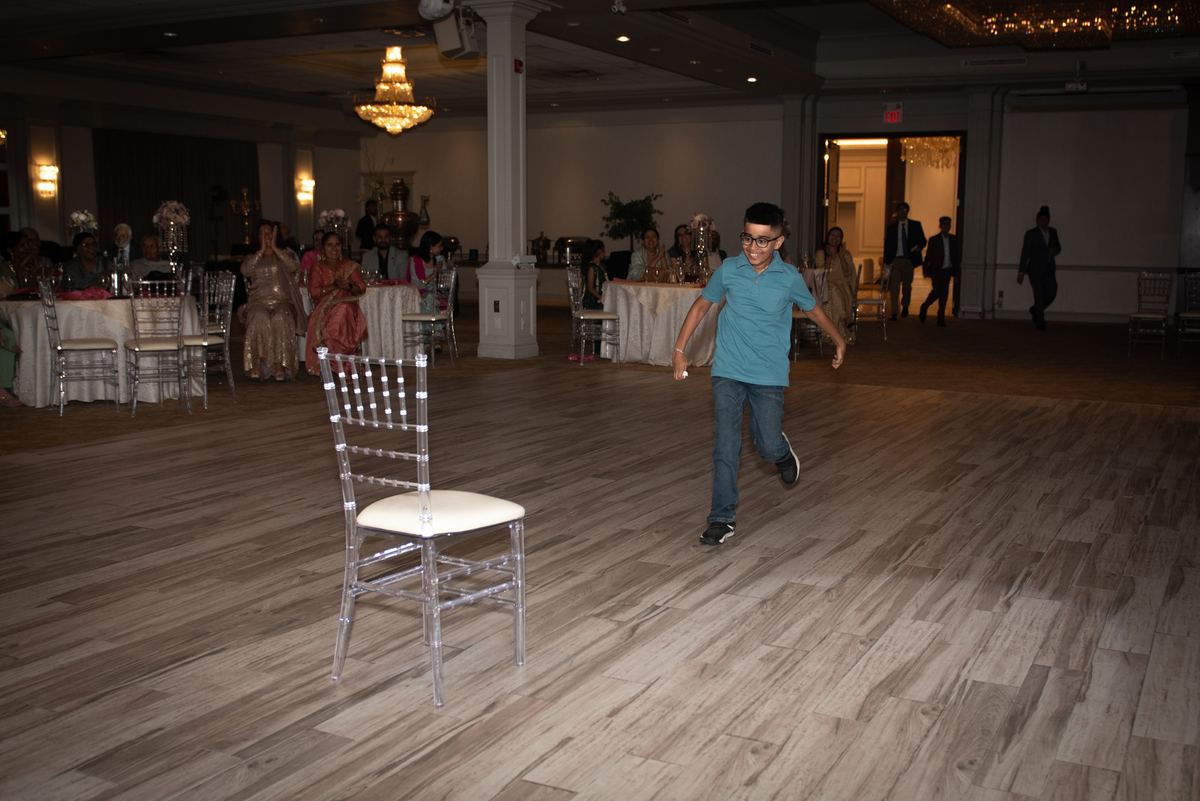 A young boy in a blue shirt runs across a banquet hall floor during a festive event with guests seated at decorated tables.