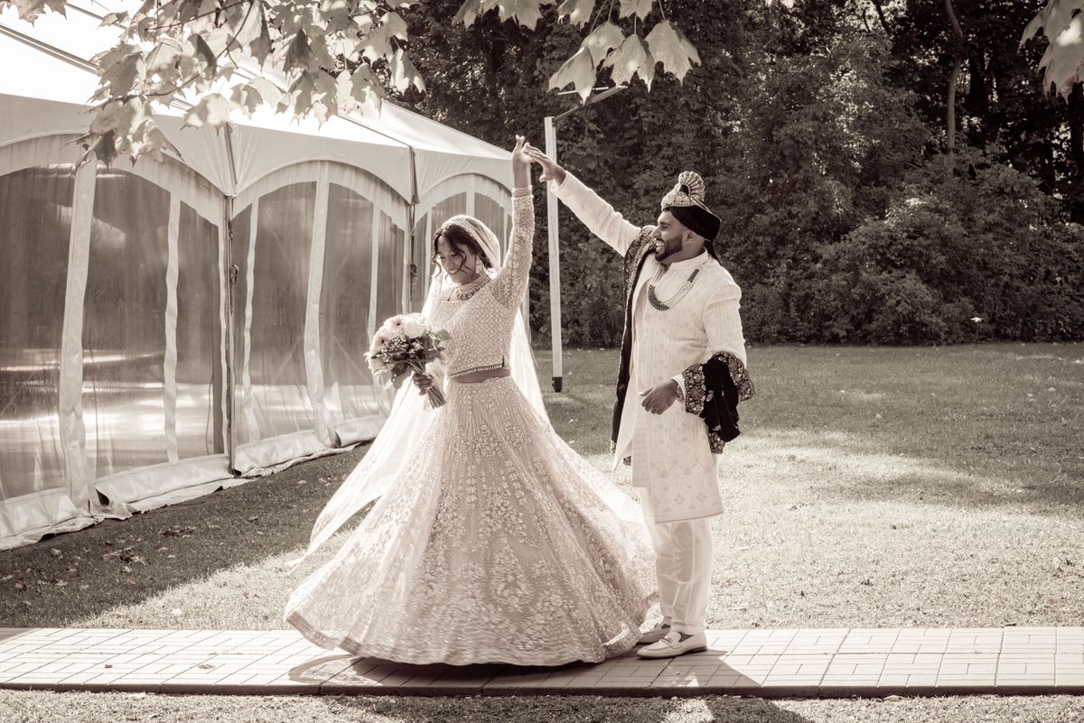 Bride in lehenga twirls as groom raises her hand near wedding tent, sunlight dapples grass and path.