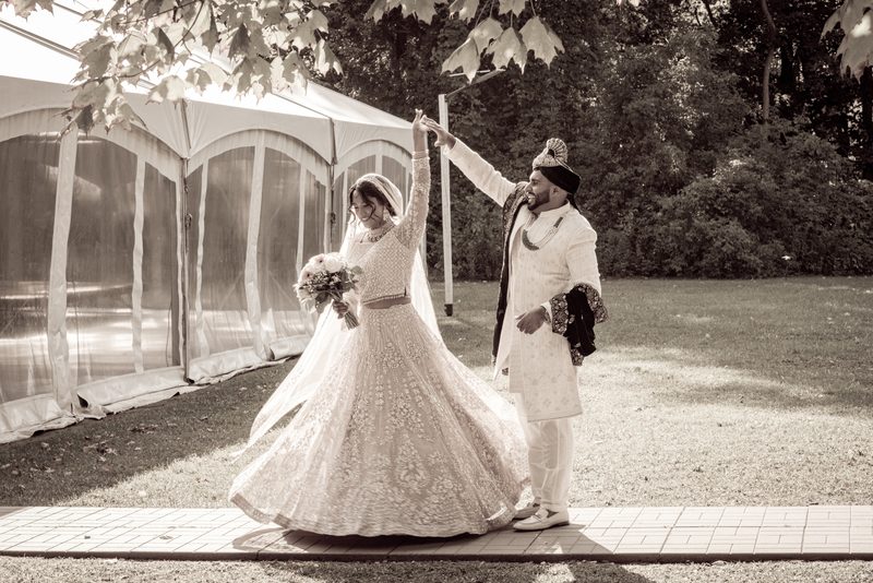 Bride in lehenga twirls as groom raises her hand near wedding tent, sunlight dapples grass and path.