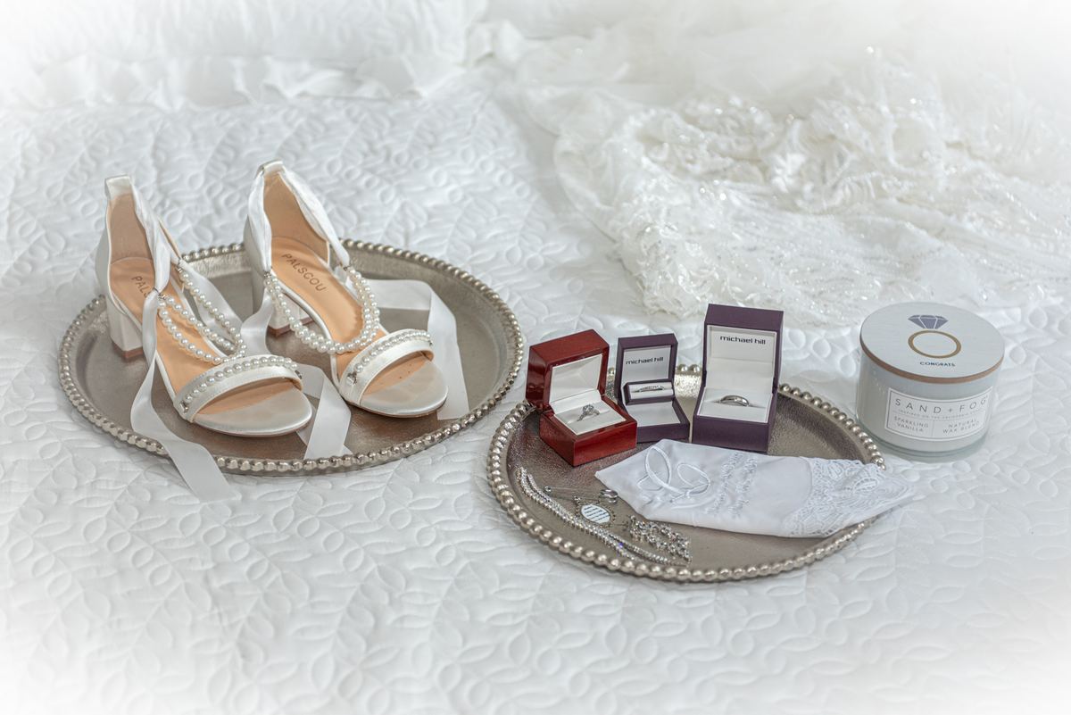 Silver trays hold wedding shoes, rings, and a lace handkerchief on a white quilted bedspread, illuminated by soft light.