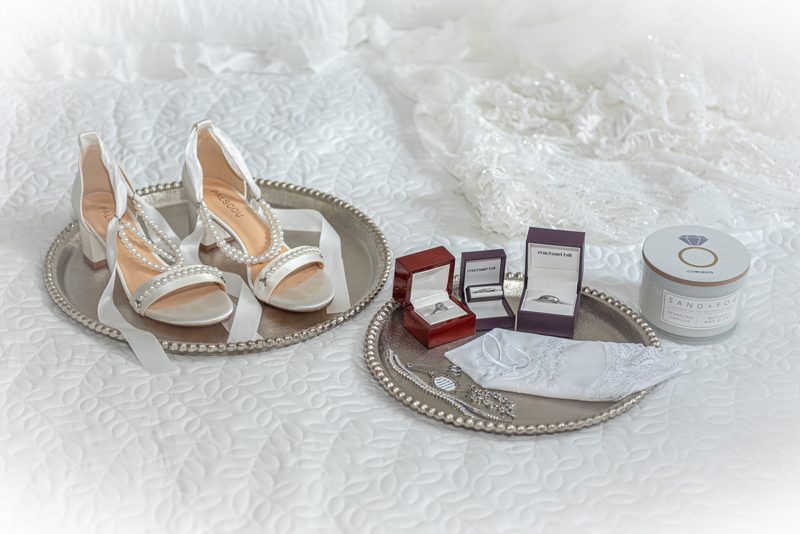 Silver trays hold wedding shoes, rings, and a lace handkerchief on a white quilted bedspread, illuminated by soft light.