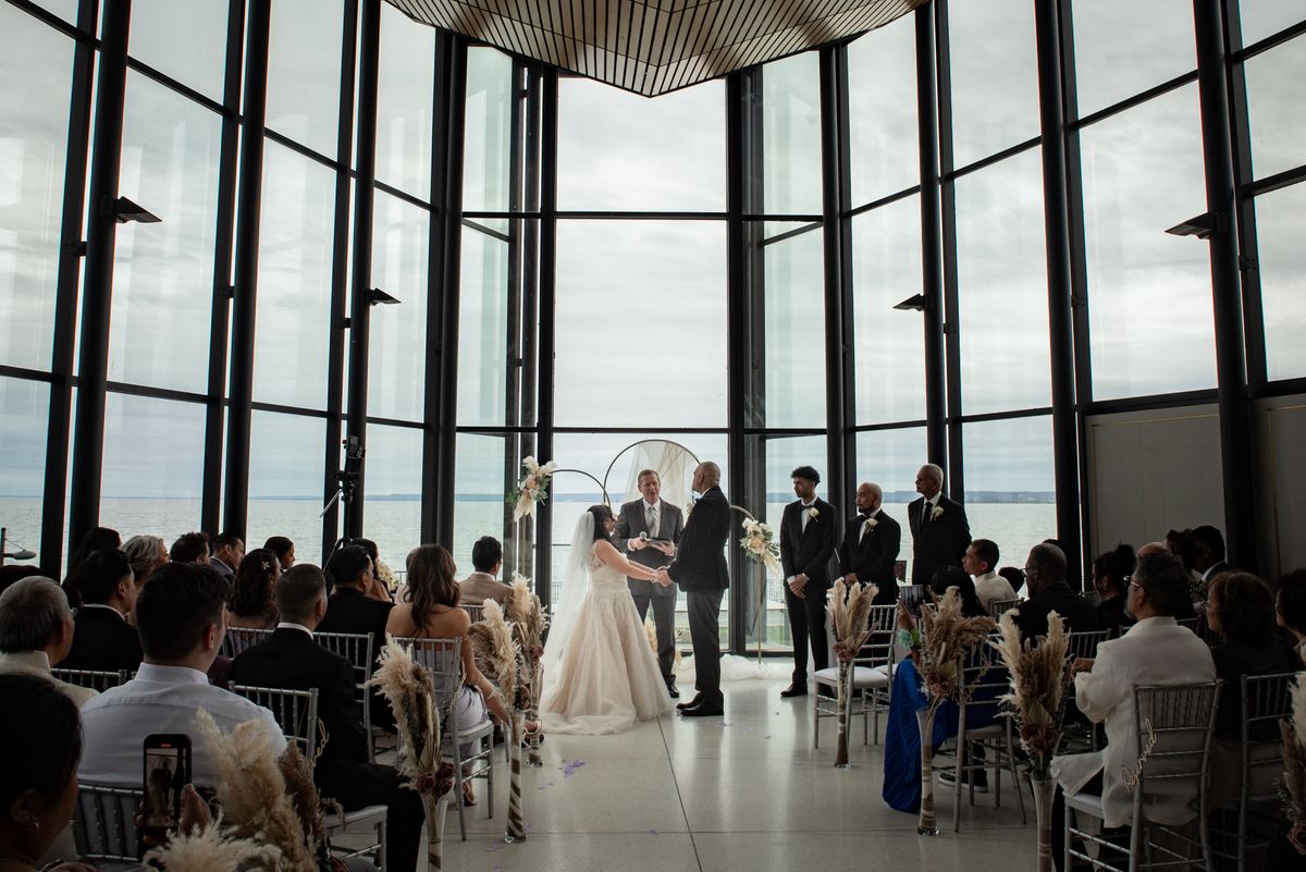 Bride and groom exchange vows at an altar with large windows overlooking water, guests watching in rows.