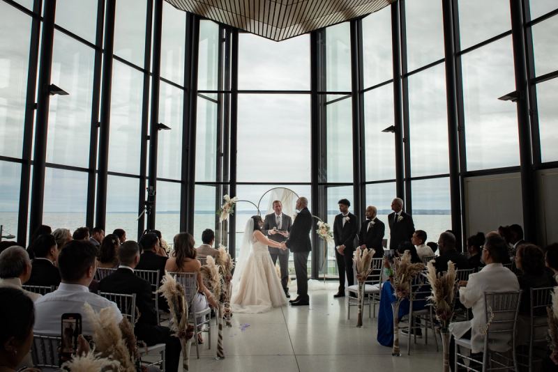 Bride and groom exchange vows at an altar with large windows overlooking water, guests watching in rows.