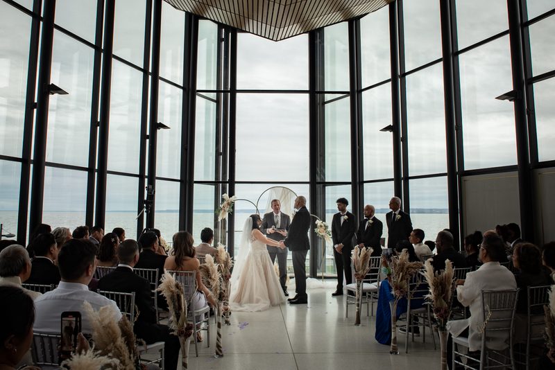 Bride and groom exchange vows at an altar with large windows overlooking water, guests watching in rows.