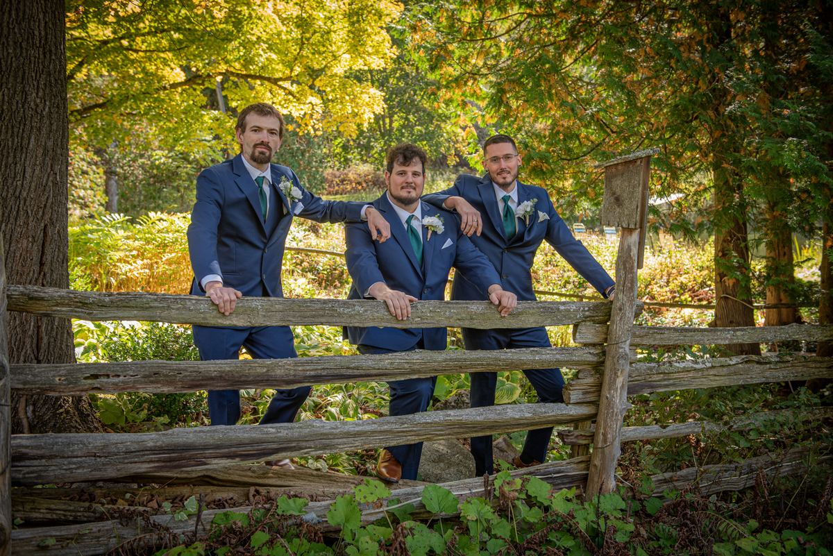 Three men in navy suits and green ties stand behind a wooden fence in an autumn forest bathed in dappled sunlight.
