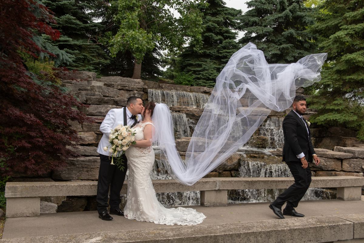 Bride and groom kiss by waterfall, framed by green and burgundy foliage, with a man in a black suit walking nearby.