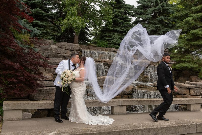 Bride and groom kiss by waterfall, framed by green and burgundy foliage, with a man in a black suit walking nearby.