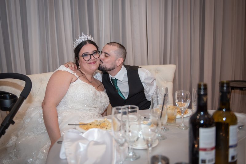 A bride in a white gown and tiara smiles as her groom kisses her cheek during a celebratory meal at their wedding reception.