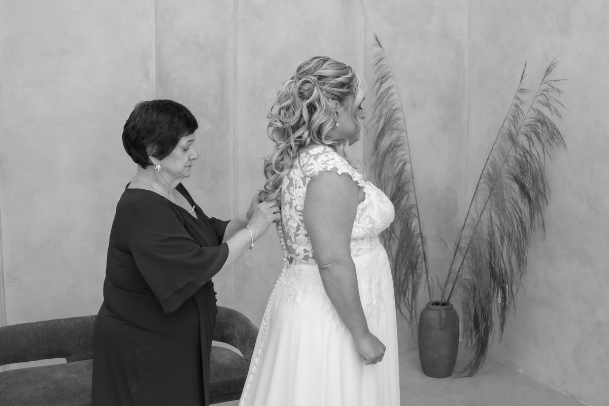 A bride's attendant adjusts the lace at the back of her wedding dress as she stands patiently near pampas grass.