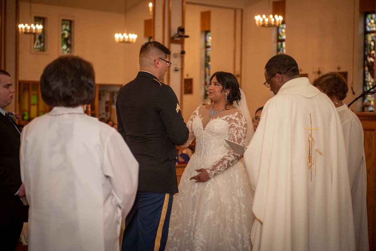 Bride in lace gown and groom in military uniform stand at altar during church wedding, illuminated by chandeliers.