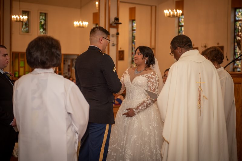 Bride in lace gown and groom in military uniform stand at altar during church wedding, illuminated by chandeliers.
