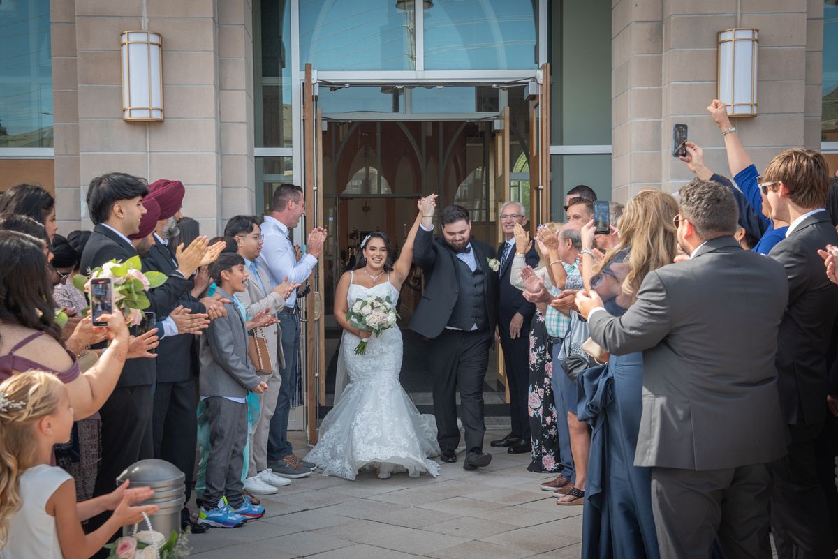 A joyful bride and groom exit a modern building with arms raised as guests cheer and clap, phones out to capture the moment.
