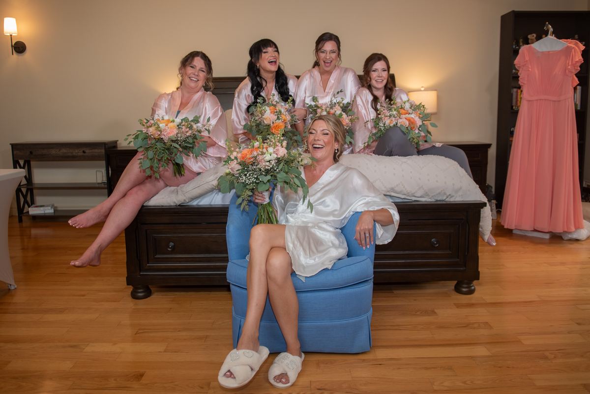 A bride and her bridesmaids in satin robes laugh together while holding flowers near a bed and chair.