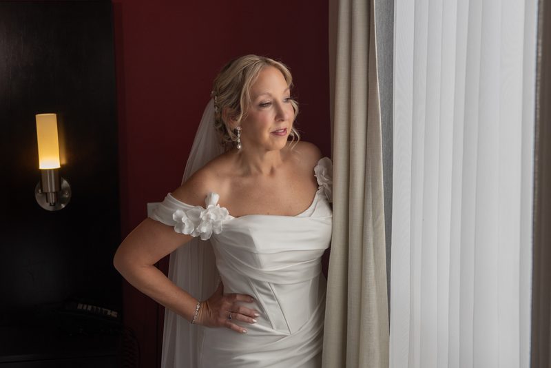 Bride in an off-the-shoulder gown with floral details stands by a window, illuminated by soft light from a sconce.