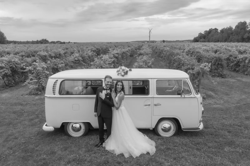 A bride and groom laugh beside a vintage white Volkswagen bus parked among the vineyard rows, captured in black and white.