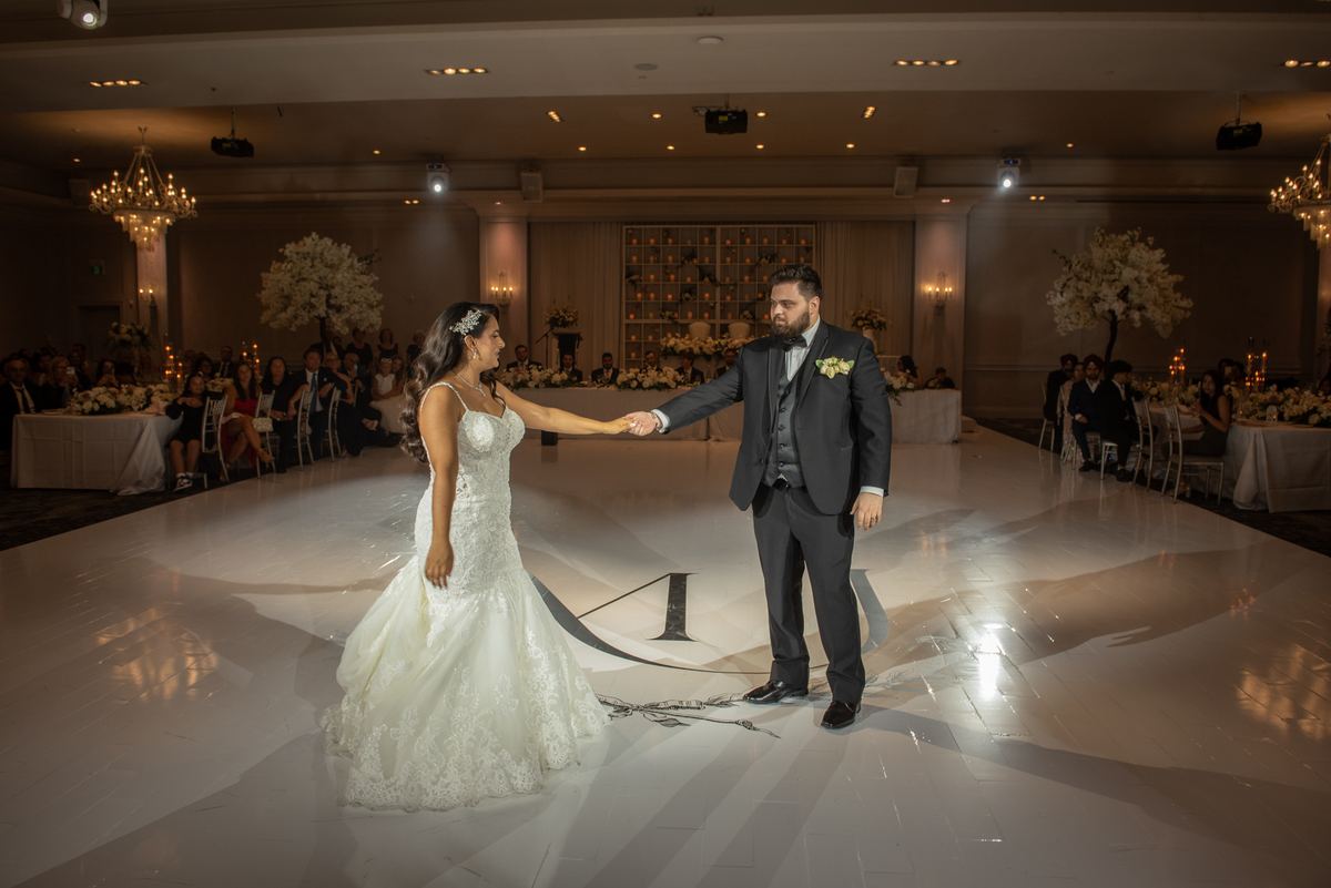 A bride in a white lace gown and groom in a dark suit hold hands while dancing at center stage during their reception.