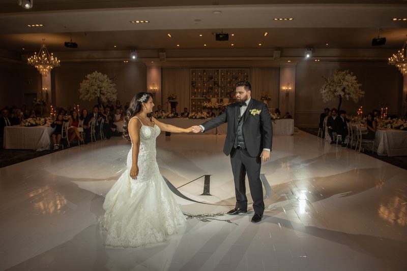 A bride in a white lace gown and groom in a dark suit hold hands while dancing at center stage during their reception.