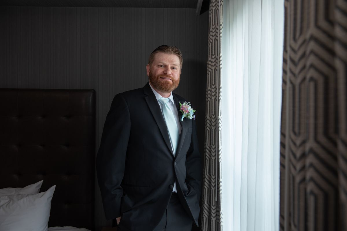 A bearded groom in a suit stands by a window with sheer curtains in a hotel room, hands in pockets, smiling.
