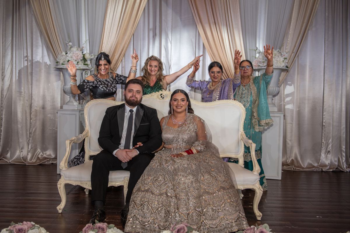 Bride in an ornate lehenga and groom in a tuxedo sit on a white settee with four women behind them, all smiling.