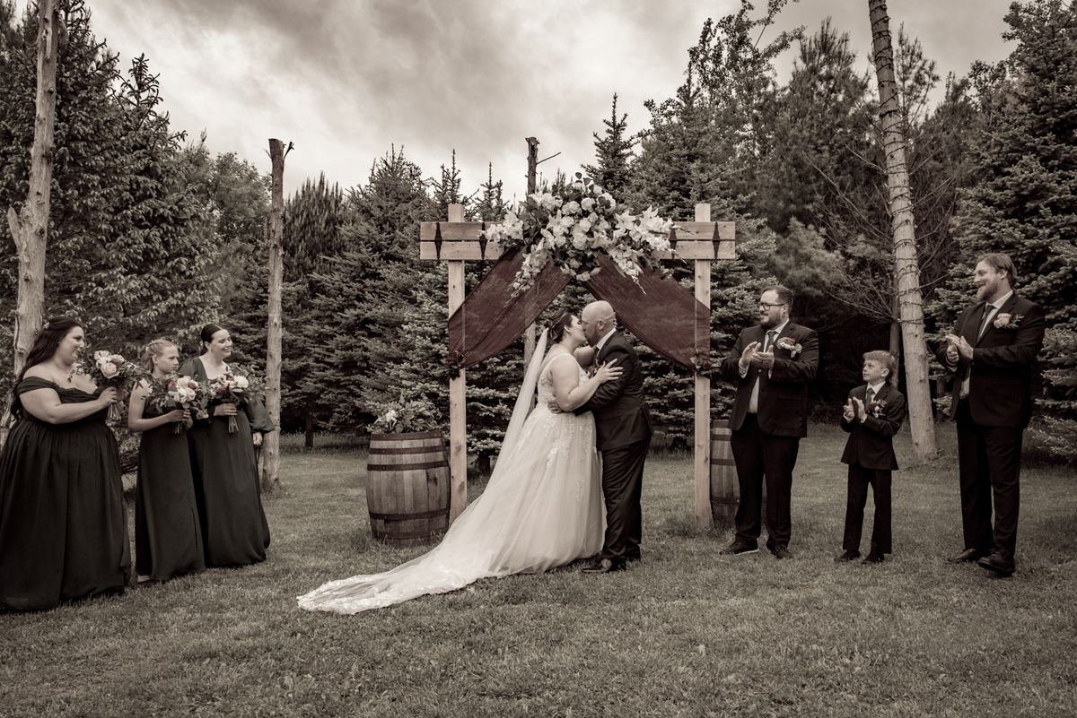 A newlywed couple kisses under a floral arch as their wedding party celebrates in a woodland setting.