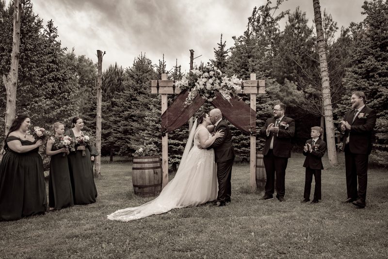 A newlywed couple kisses under a floral arch as their wedding party celebrates in a woodland setting.