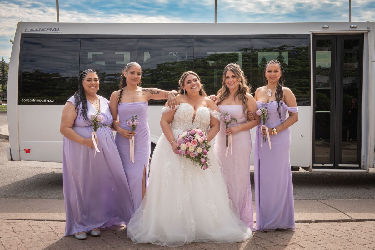 A bride and four bridesmaids pose joyfully in front of a white limousine, holding bouquets on a sunny day.
