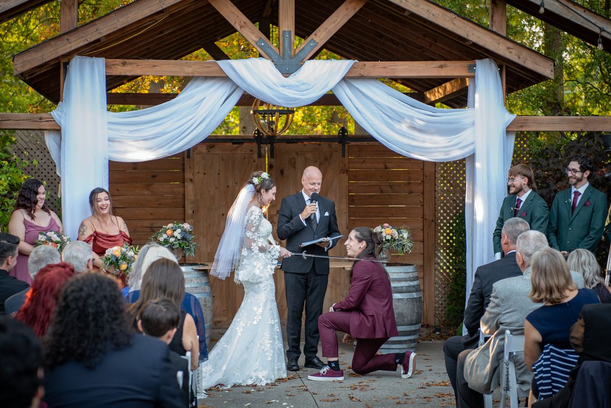 A bride in a lace gown and veil stands beside her kneeling groom under a wooden arbor, surrounded by guests.