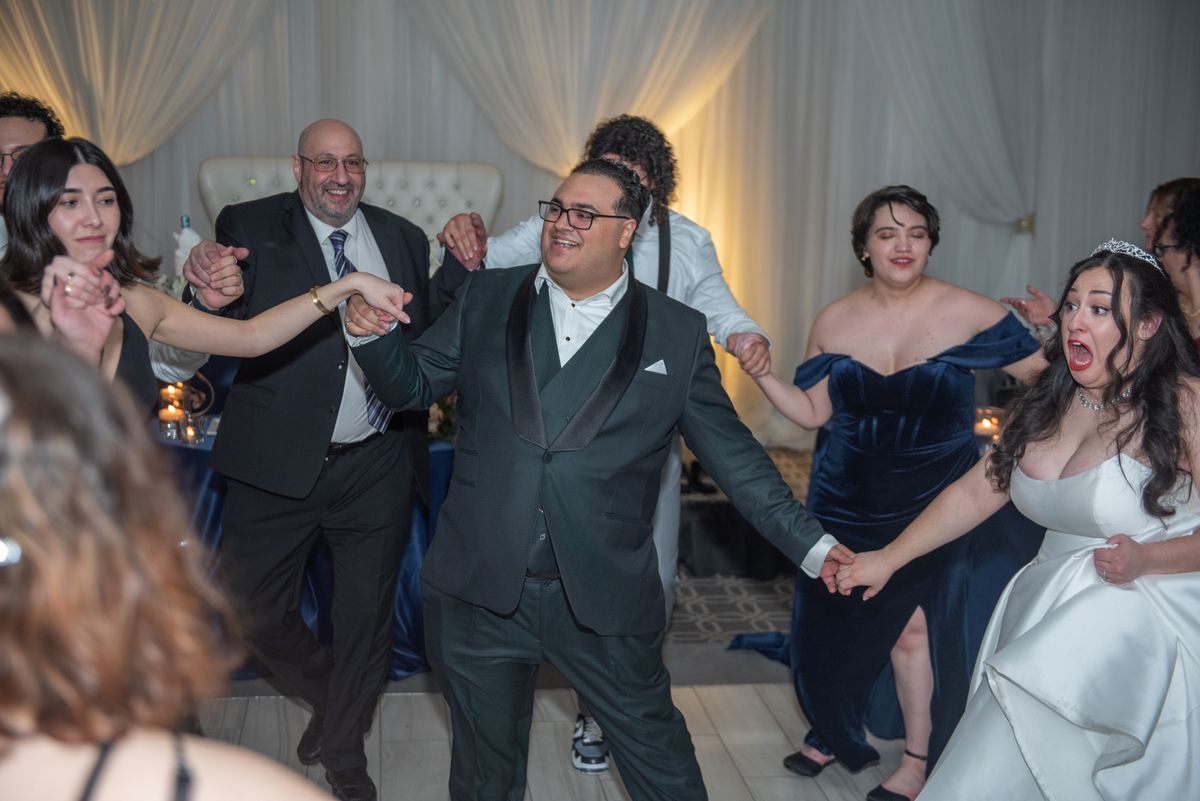 Wedding guests, including the bride and groom, dance energetically in a circle under warm ambient lighting from sheer drapes.