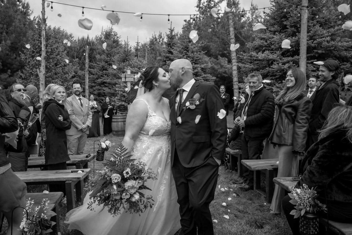 Anastasia and Adam share a tender kiss at their outdoor wedding ceremony, surrounded by guests seated on wooden benches.