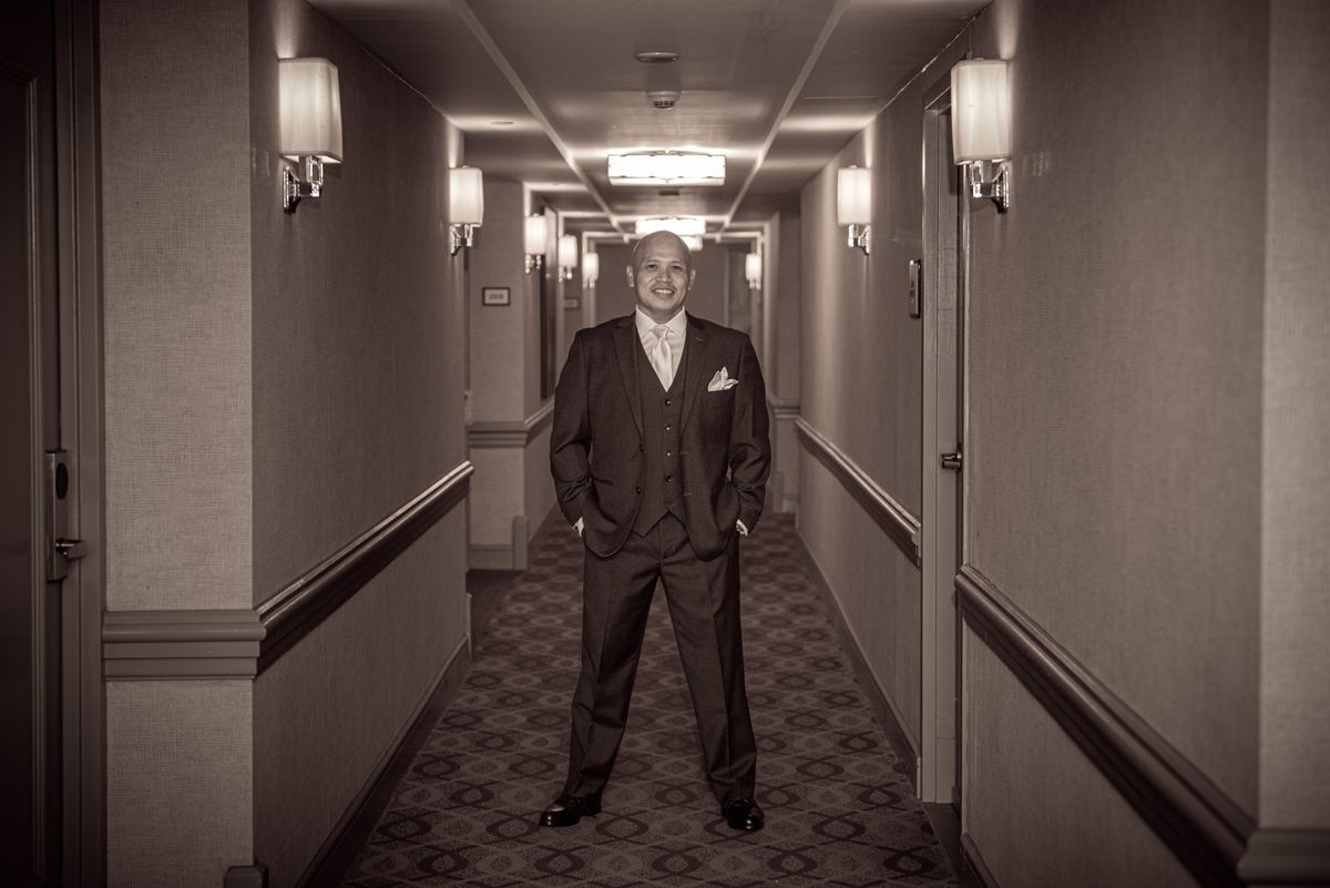 A groom in formal attire stands confidently in a hotel corridor bathed in soft, warm light from wall sconces.