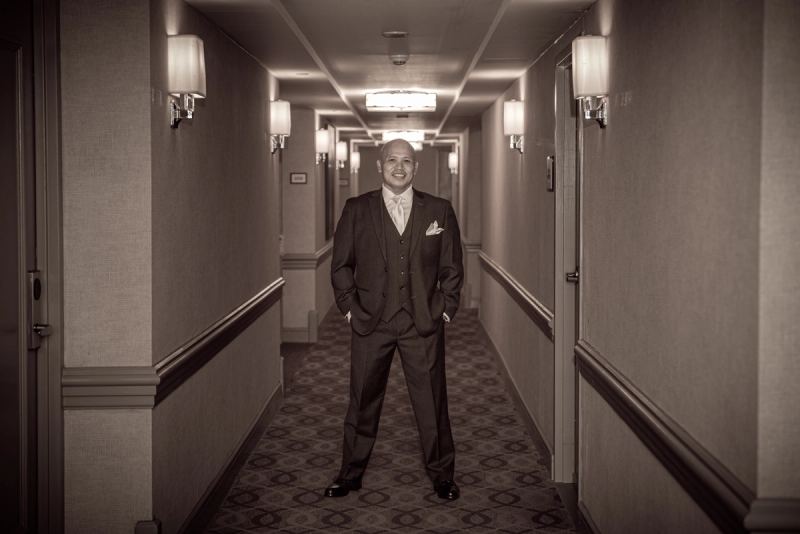 A groom in formal attire stands confidently in a hotel corridor bathed in soft, warm light from wall sconces.