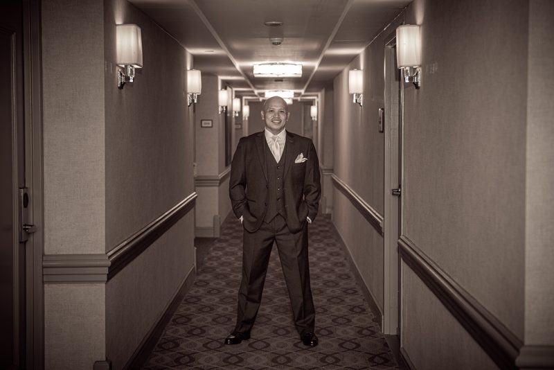 A groom in formal attire stands confidently in a hotel corridor bathed in soft, warm light from wall sconces.