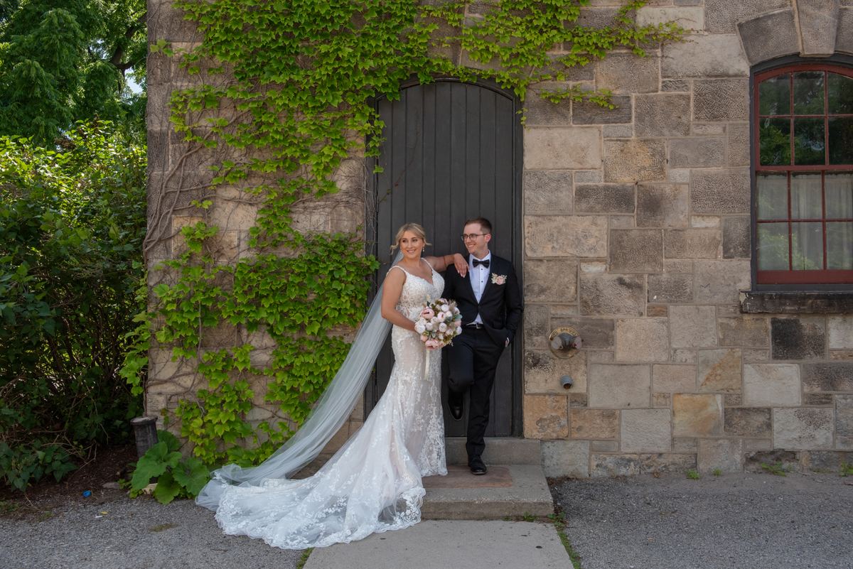 A bride and groom pose joyfully by a stone doorway covered in green ivy, illuminated by soft natural light.