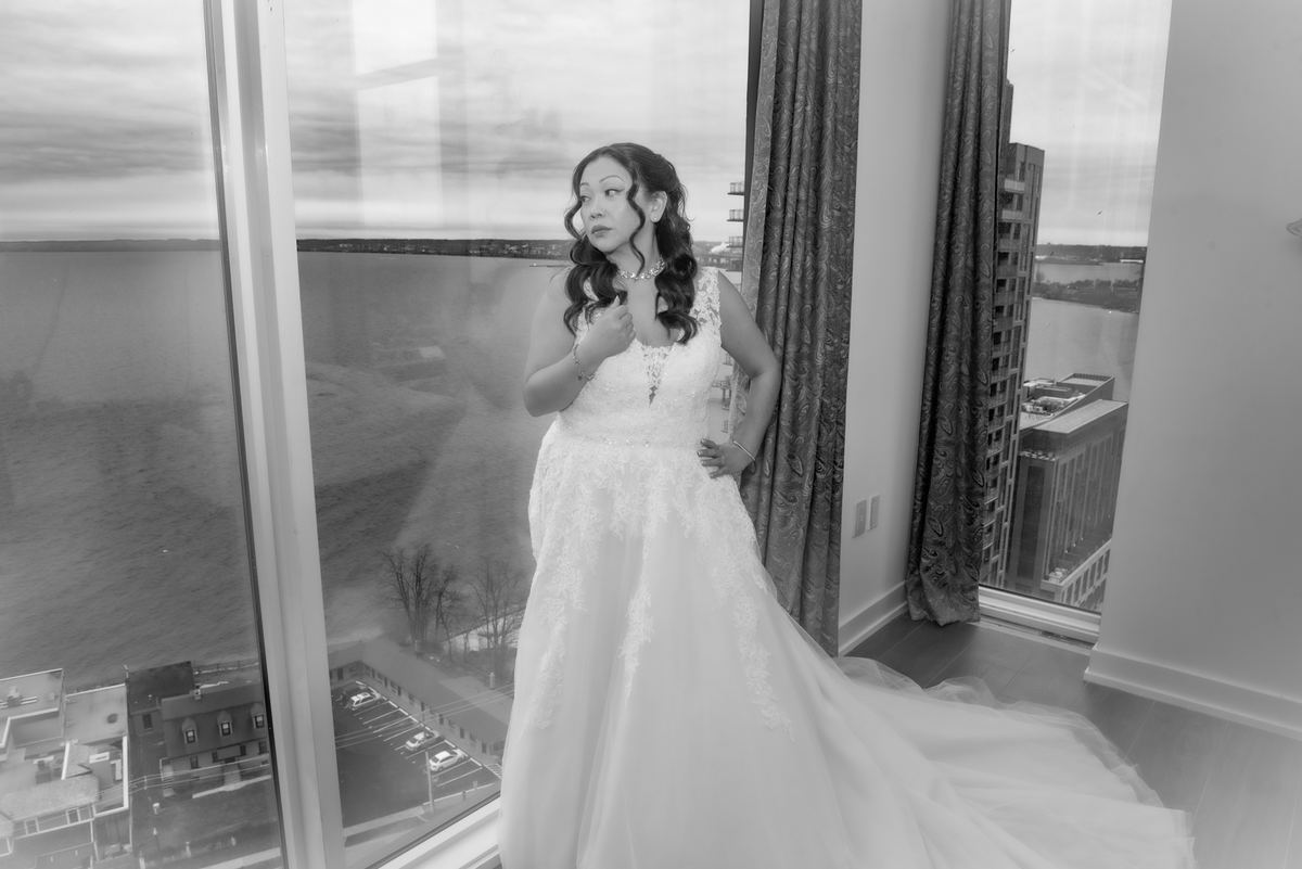 A bride in a lace gown stands by a window, gazing at a waterfront cityscape, her reflection visible in the glass.