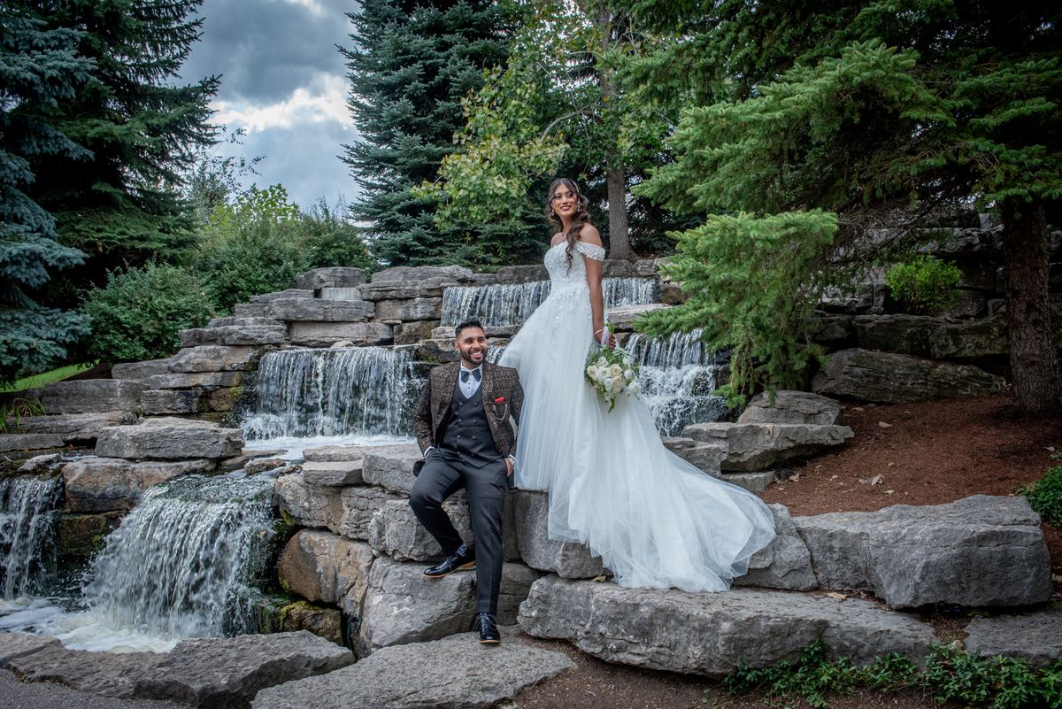A bride in a white gown and groom sit by a stone waterfall in a forested garden, both smiling warmly under an overcast sky.