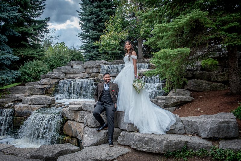 A bride in a white gown and groom sit by a stone waterfall in a forested garden, both smiling warmly under an overcast sky.
