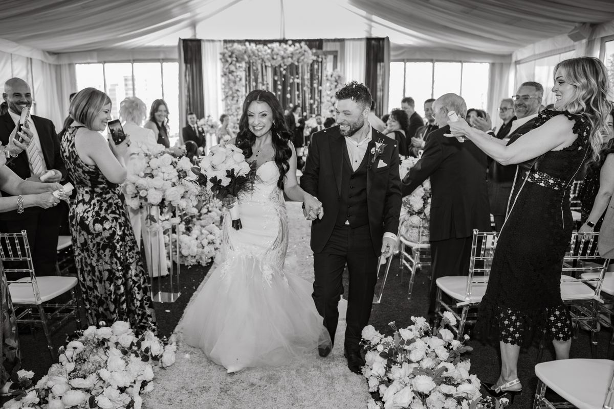 A smiling bride and groom walk down the aisle of a tented wedding space, surrounded by cheering guests holding phones.