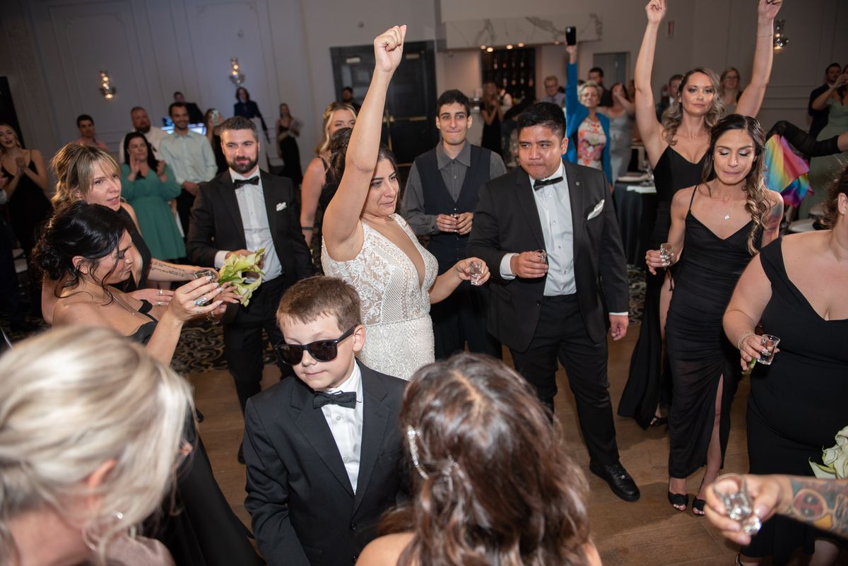 A woman in a white lace gown raises her fist among dancing guests in formal attire, illuminated by warm lights.