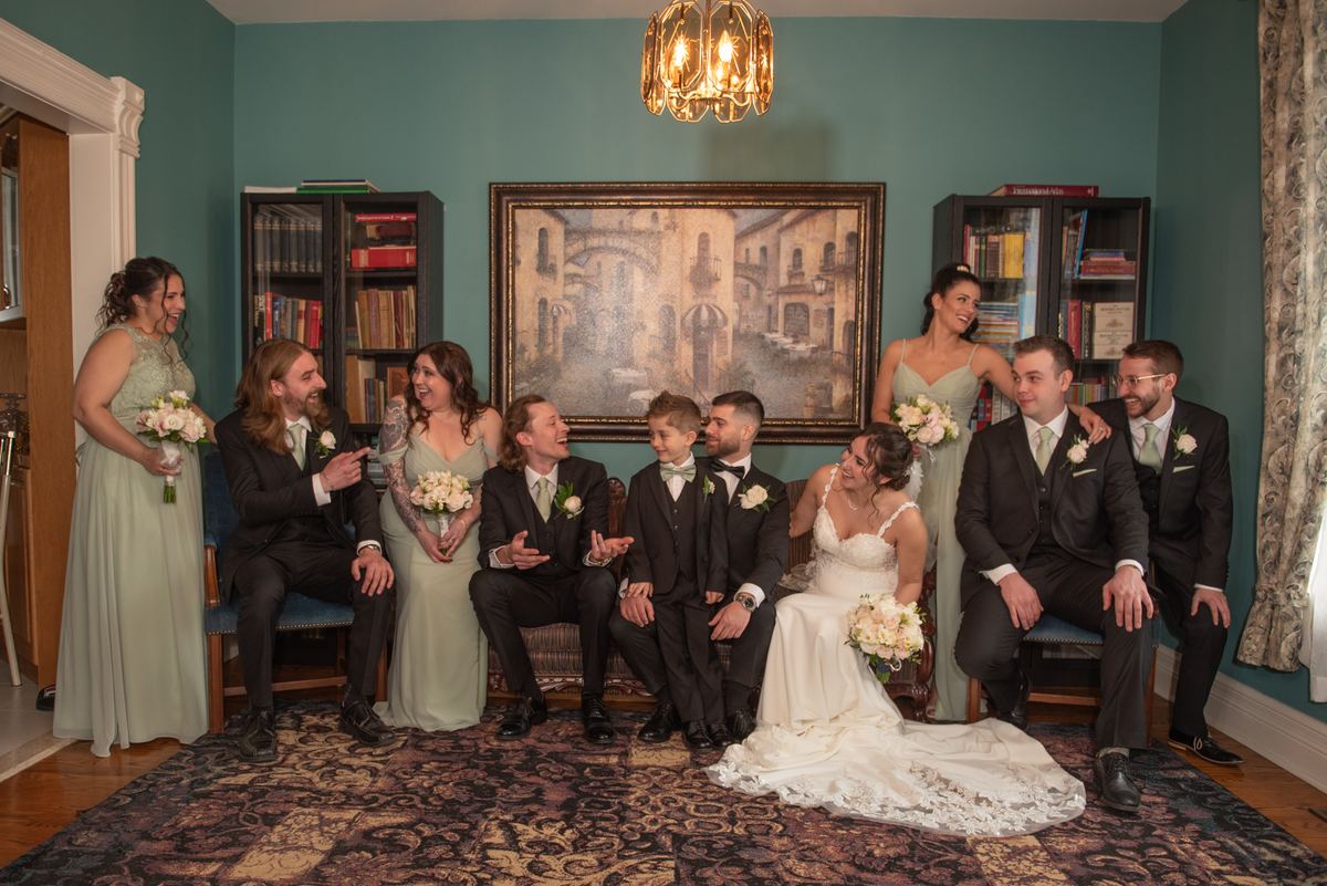 Wedding party celebrates in a warmly lit room with teal walls and bookshelves, surrounded by bridesmaids and groomsmen.