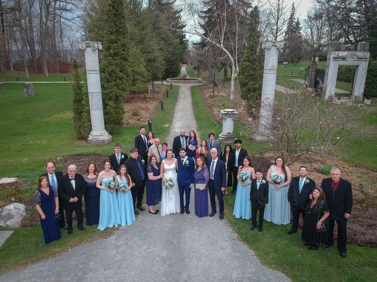 Bride and groom stand with wedding party and family along a gravel path bordered by stone columns and greenery.