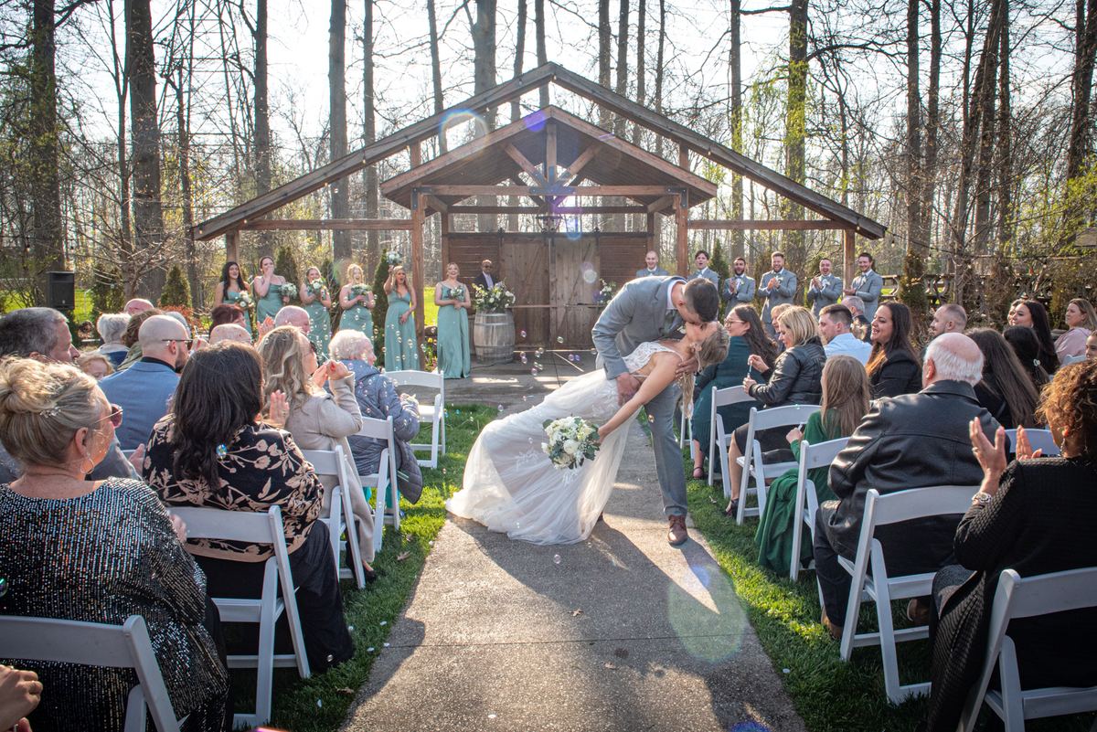 A bride and groom kiss at an altar as guests applaud, petals scatter, and sunlight filters through trees.