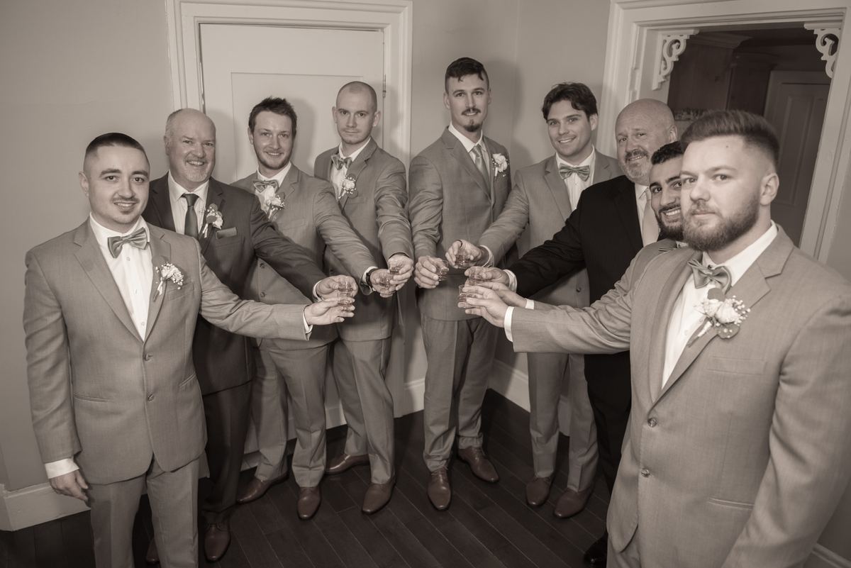 Groomsmen and the groom in formal attire toasting with glasses, smiling and focused under soft indoor lighting.