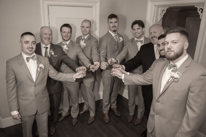 Groomsmen and the groom in formal attire toasting with glasses, smiling and focused under soft indoor lighting.